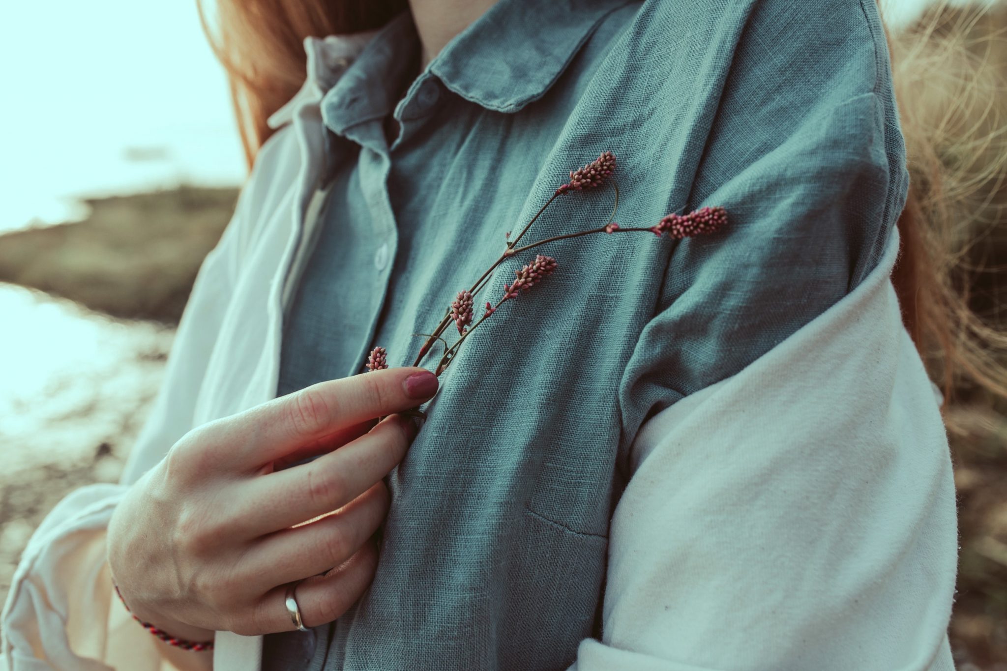 A close-up of woman's hand holding a wild flower. A young ginger woman standing by the lake shore in summertime. A red headed girl wearing i linen aquamarine blouse and linen white jacket.